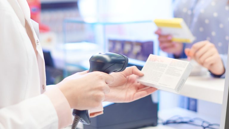 E-Rezept einlösen: Eine Apothekenmitarbeiterin steht am Verkaufstresen und scannt eine Medizinpackung, im Hintergrund steht eine Kundin am Tresen.