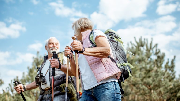 Das Bild zeigt ein gut gelauntes älteres Paar, das bei schönem Wetter mit Nordic Walking Stöcken und Outdoor -Ausrüstung durch die Natur spaziert und offensichtlich die Gemeinschaft des anderen genießt.