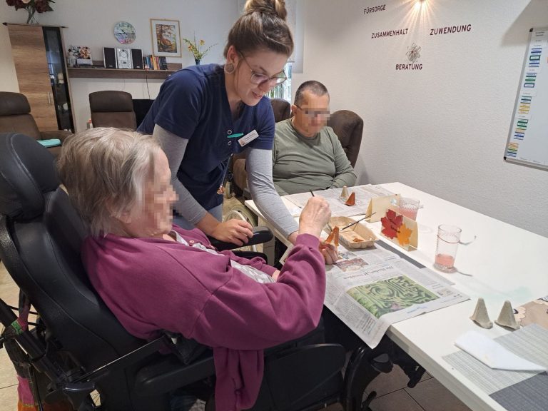 Zwei Senioren und eine Pflegerin sitzen bzw. stehen an einem Tisch, vor sich Bastelmaterialien. Sie agieren mit beim Herbstbasteln in der solitas Tagespflege.