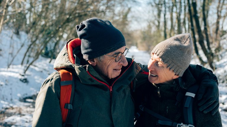 Das Bild zeigt ein älteres Pärchen. Sie laufen durch eine Winterlandschaft, halten sich dabei im Arm und lächeln sich an. Gesund und fit durch den Winter zu kommen, ist besonders auch für ältere Menschen essentiell.