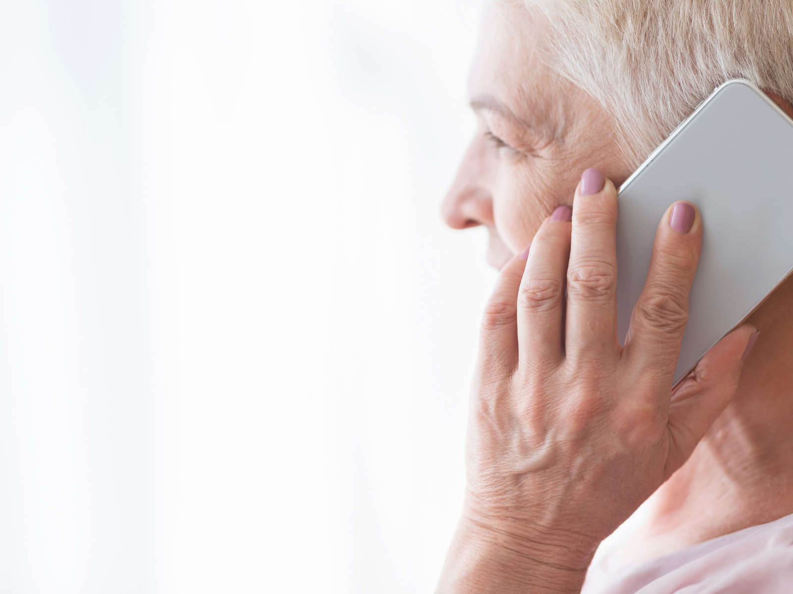 Das Bild zum Beitrag über den Enkeltrick zeigt eine ältere Dame mit grauweißen Haaren im Profil, die sich mit der linken Hand ein weißes Smartphone ans Ohr hält.
