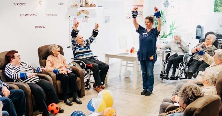 Das Bild zeigt die Räume der solitas Tagespflege. Senioren sitzen in einem Kreis und führen gemeinsam mit einer Pflegekraft schonende Fitness-Übungen durch.