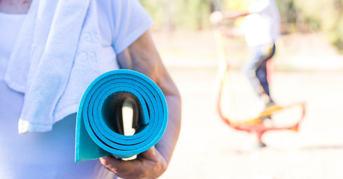 Sport für Senioren: Eine ältere Frau hält eine blaue zusammengerollte Gymnastikmatte im Arm, im Hintegrund trainiert eine Person auf einem Outdoor-Sportgerät.