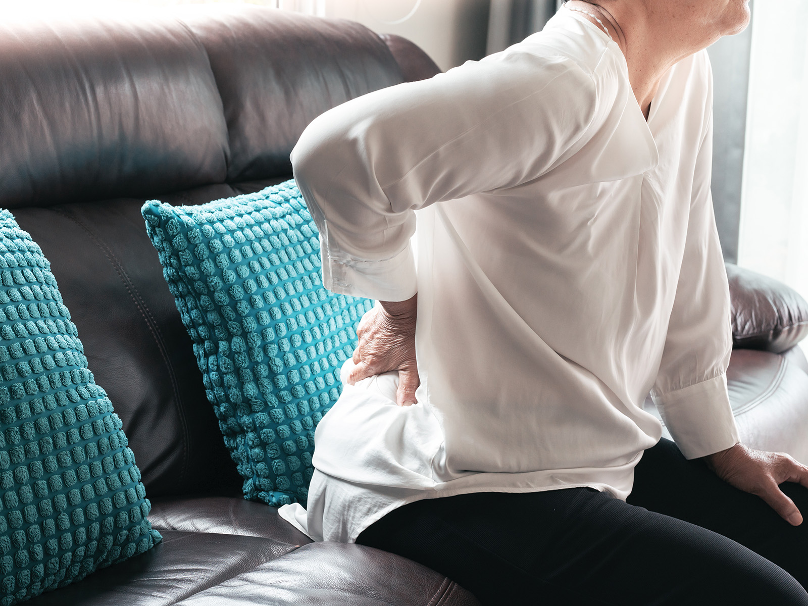 Eine Frau sitzt auf einem Sofa und hält die rechte Hand auf ihr schmerzende Lende. Gürtelrose kann starke Schmerzen im Rückenbereich und anderen Körperregionen verursachen.