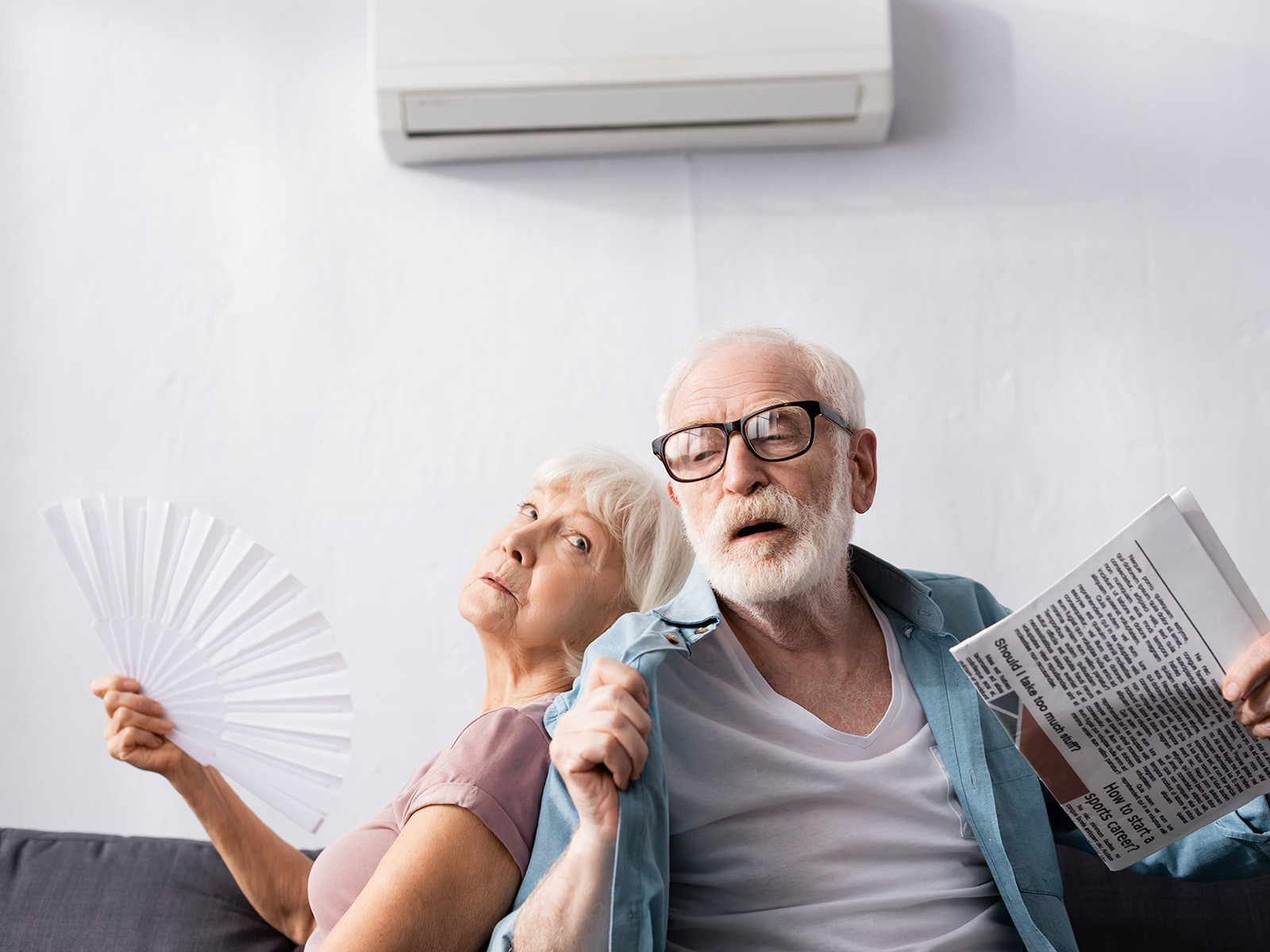 Umgang mit Wetterkapriolen, Schwüle und Hitze: Älterer Mann hält Zeitung neben müder Frau mit Ventilator unter Klimaanlage im Wohnzimmer