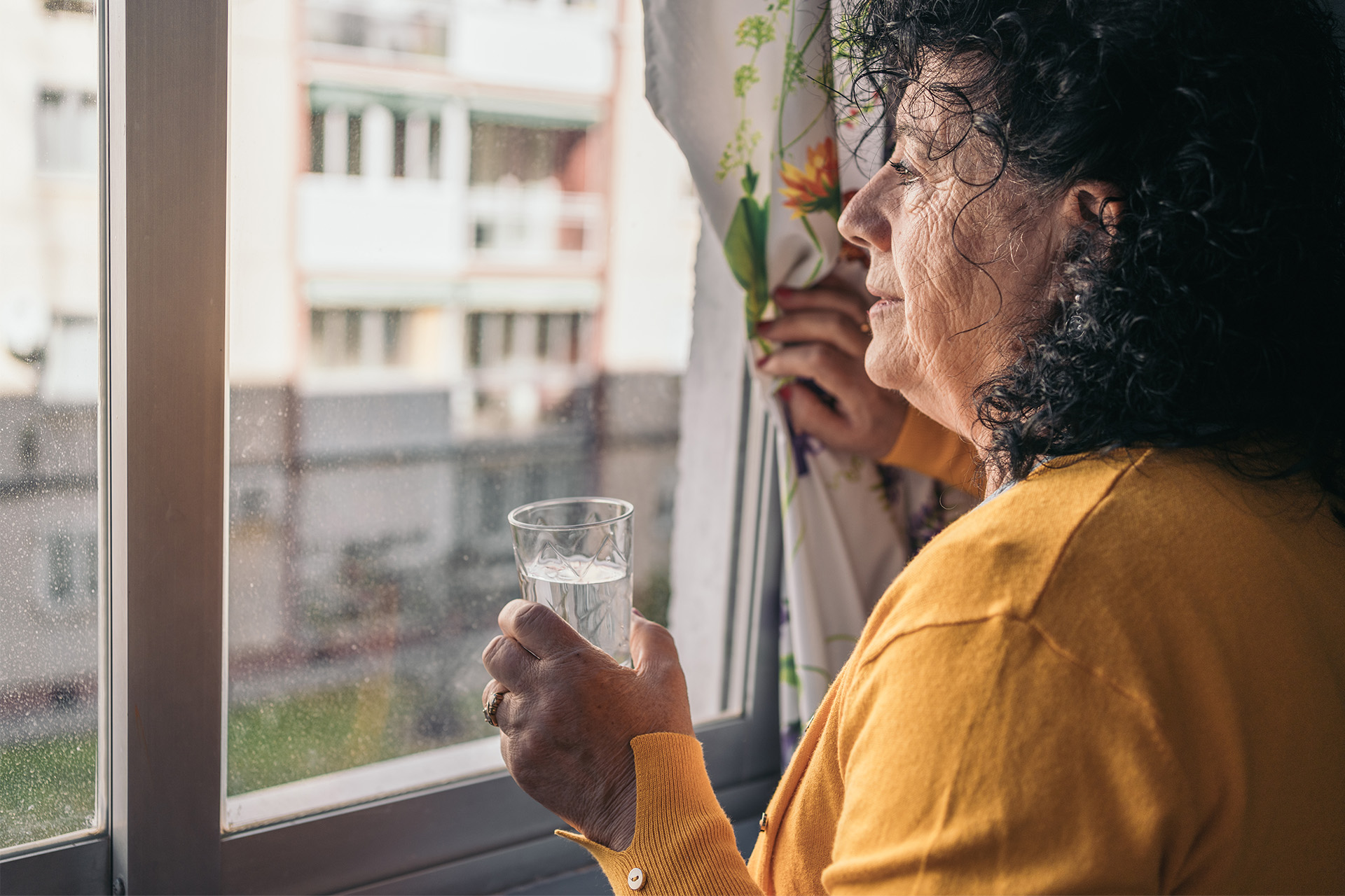 Ältere Frau, die aus dem Fenster schaut, während sie mit einer Hand den Vorhang zurückzieht und mit der anderen ein Glas Wasser hält.