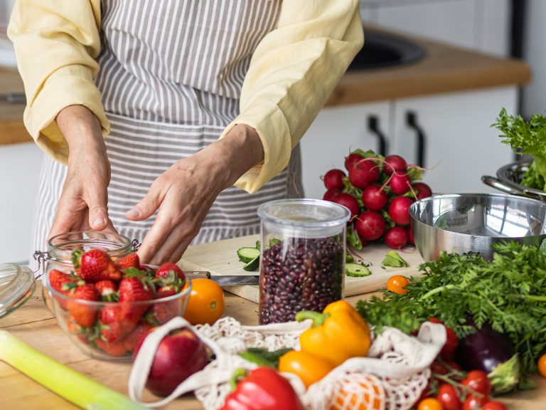 Ältere Dame steht in einer Küche. Auf der Arbeitsplatte vor sich stehen zahlreiche gesunde Zutaten für eine sommerliche Ernährung, darunter Tomaten, Paprika, Radieschen und Kidneybohnen.