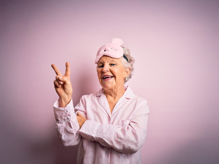 Besser schalfen im Alter - schöne ältere Frau mit rosa Schlafmaske und Pyjama vor rosa Hintergrund, die mit fröhlichem Gesicht in die Kamera lächelt, zwinkert und ein Victory-Zeichen macht.