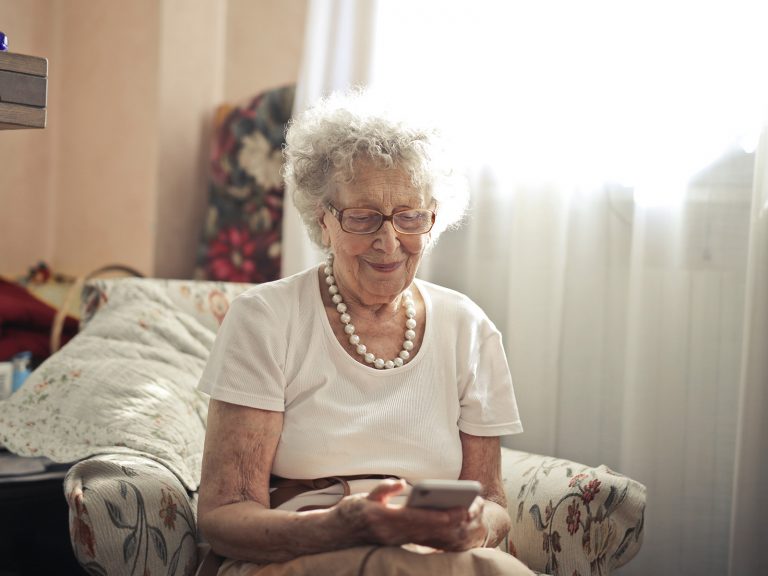 Eine Seniorin sitzt im Wohnzimmer und sieht lächelnd auf das Smartphone in ihren Händen. Digitale Gesundheitstools können insbesondere im Alter eine nützliche Hilfe sein.