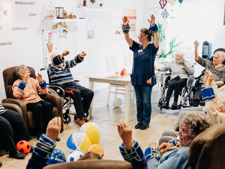 Gymnastik im Sitzen in der Tagespflege von solitas: Mehrere Senioren sitzen im Kreis, vor ihnen steht eine Pflegekraft und führt Gymnastikübrungen durch.