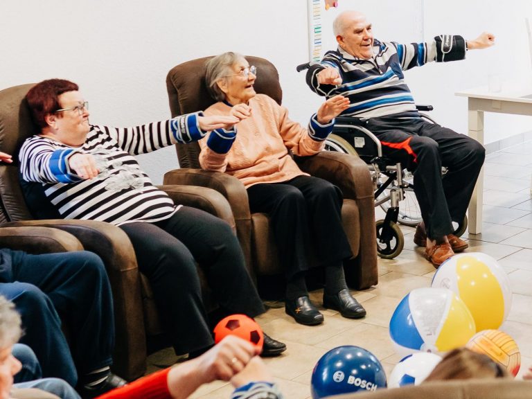 Mehrere Senioren machen Bewegungstherapie in der Tagespflege von solitas. Sie sitzen auf Sesseln und heben die Arme mit Gewichtsmanschetten. Seit der Kooperation mit Physio M6 ist das Angebot ausgeweitet worden.