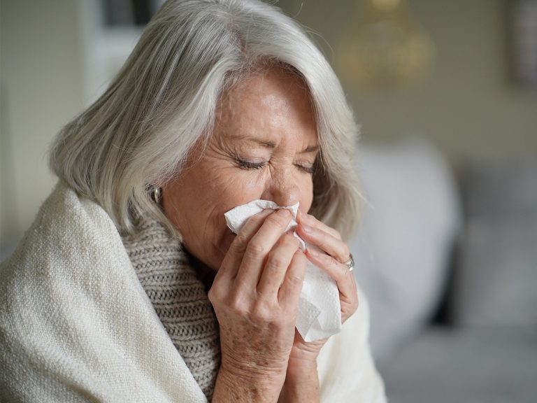 Eine ältere Dame ist in einen Schal und eine Decke gehüllt und schnaubt sich die Nase.
