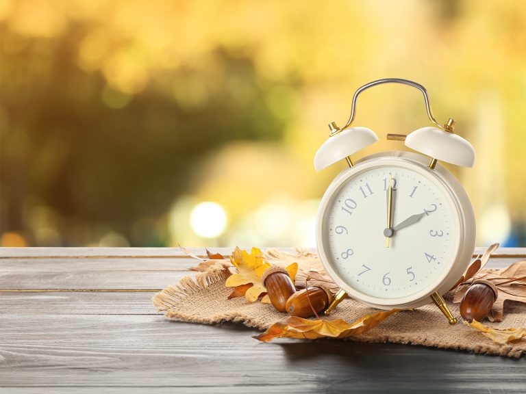 Wecker, Eicheln und Herbstblätter auf einem Tisch im Freien. Zeitumstellung