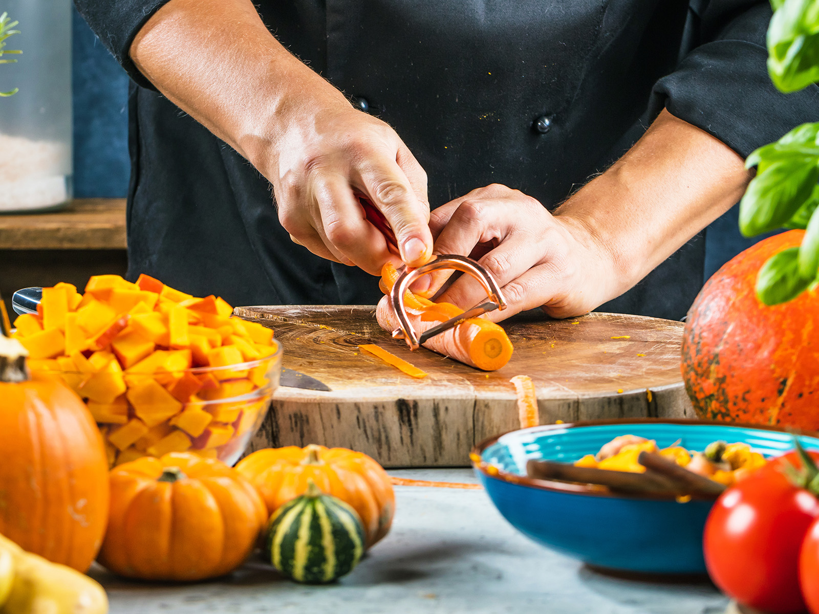 Eine Person schält auf einem Holzbrett eine Möhre, rundherum liegen Kürbisse und andere Lebensmittel