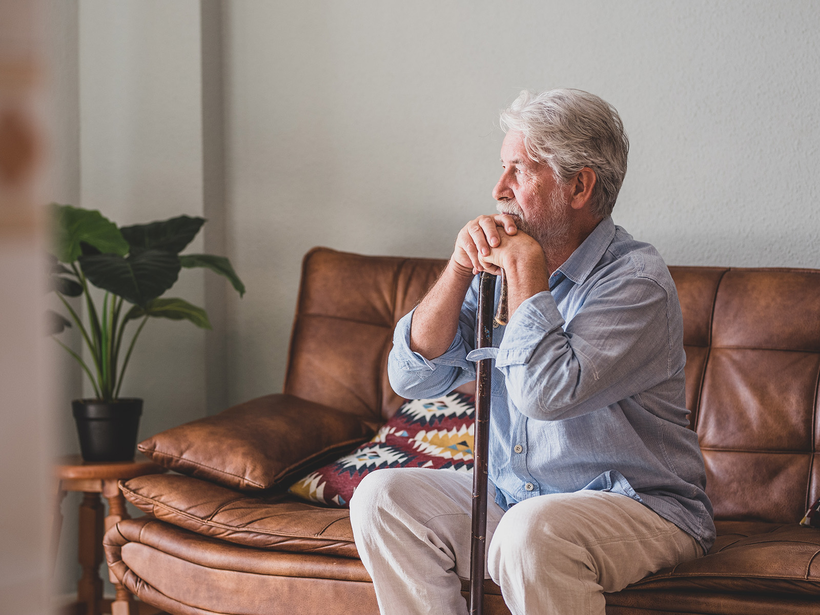 Ein älterer Mann sitzt nachdenklich auf einem braunen Sofa, die Hände und das Gesicht auf einen Stock gestützt