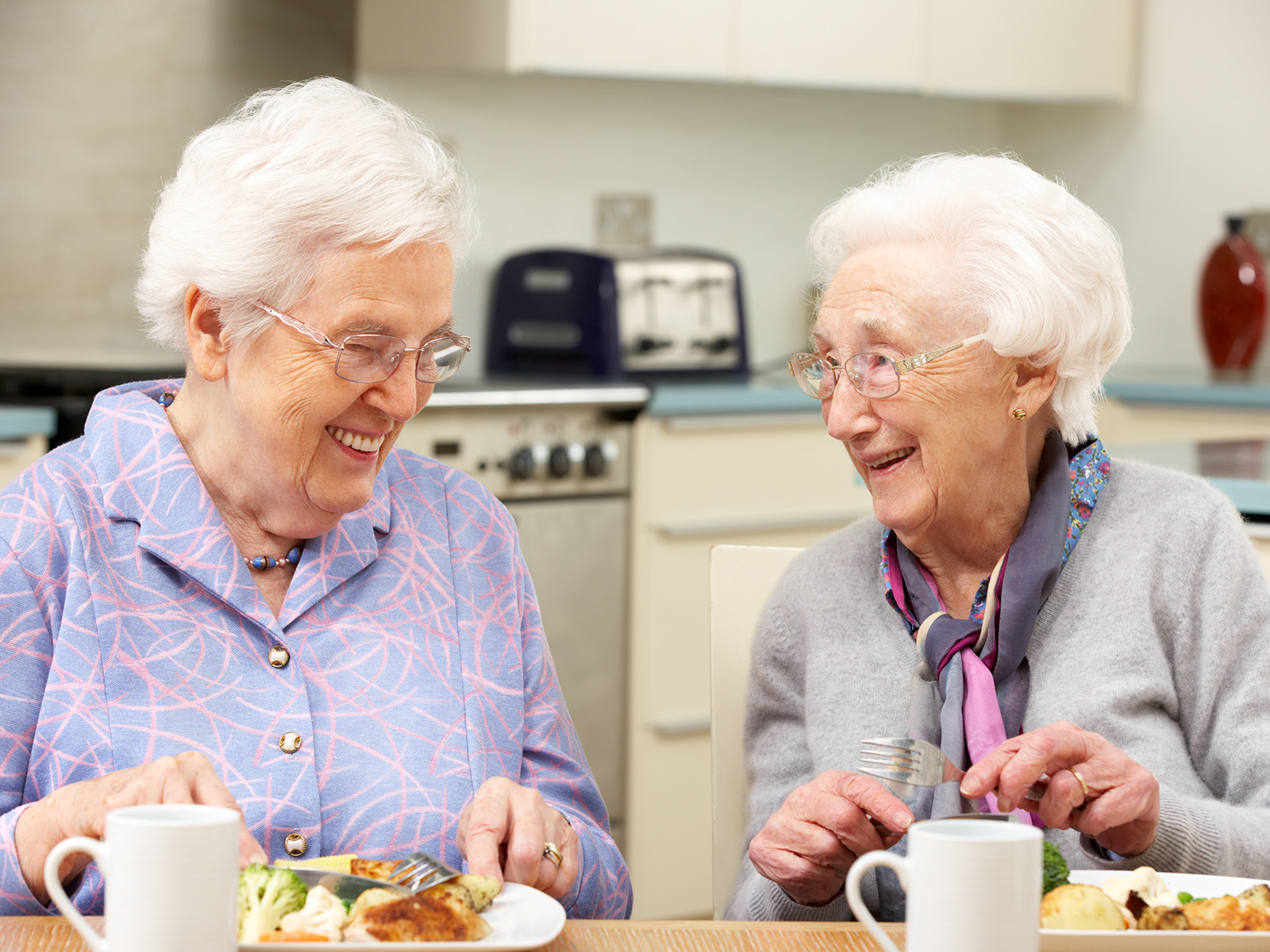 Zwei ältere Frauen sitzen essend nebeneinander. Ihre Teller sind ein Beispiel für gesunde Ernährung - Gemüse und Hühnchen.