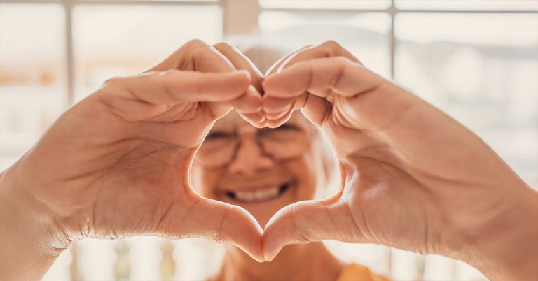 Eine Seniorin hält lachend die Hände vor die Kamera, die ein Herz formen