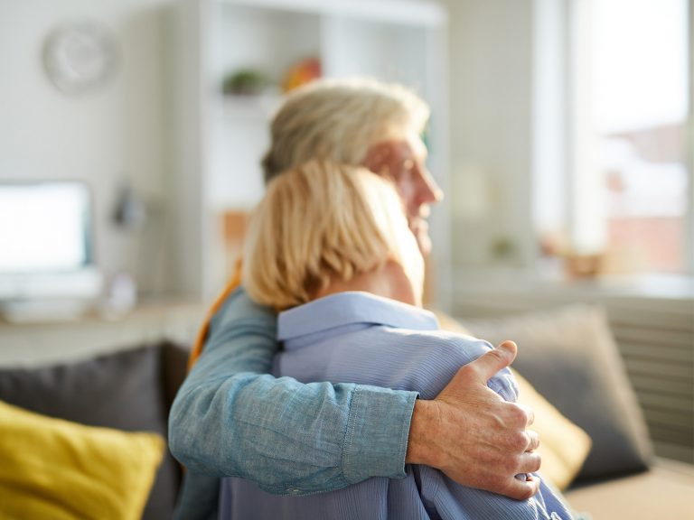 Älteres Paar sitz in Wohnzimmer, er hält sie im Arm