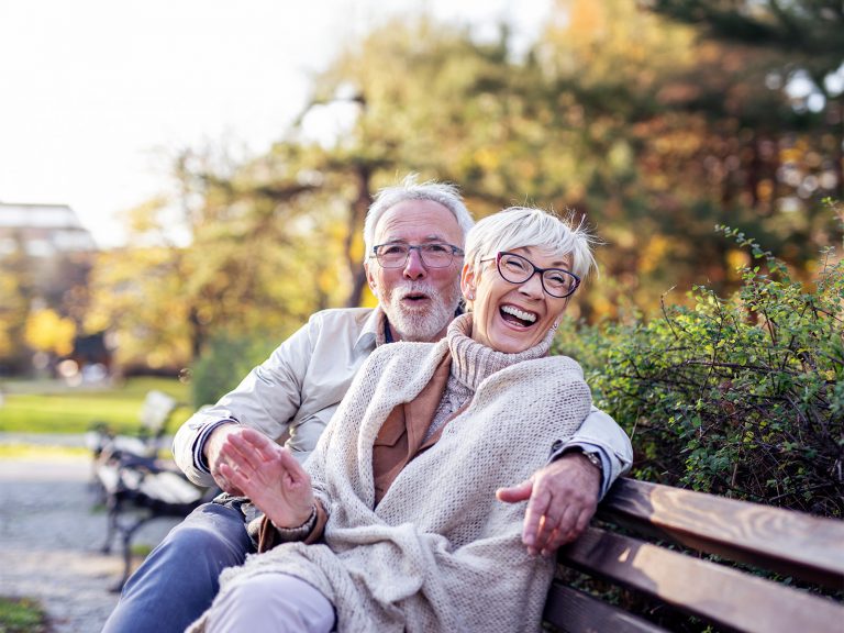 Älteres Paar sitzt auf einer Bank, er hält sie im Arm und beide lachen.