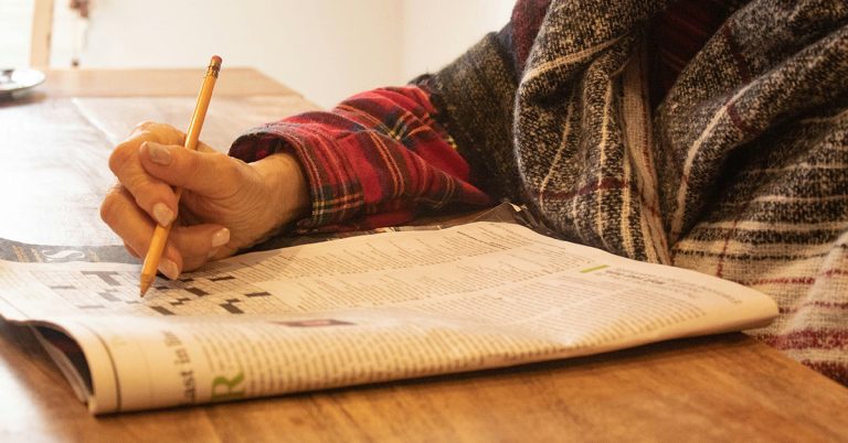 eine ältere Person sitzt im Wollpulli am Tisch und löst ein Rätsel in einer Zeitung