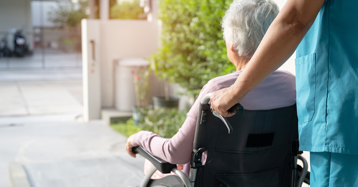 Eine Pflegeperson in blauer Klinikkleidung schiebt einen Rollstuhl, in dem eine ältere Dame sitzt