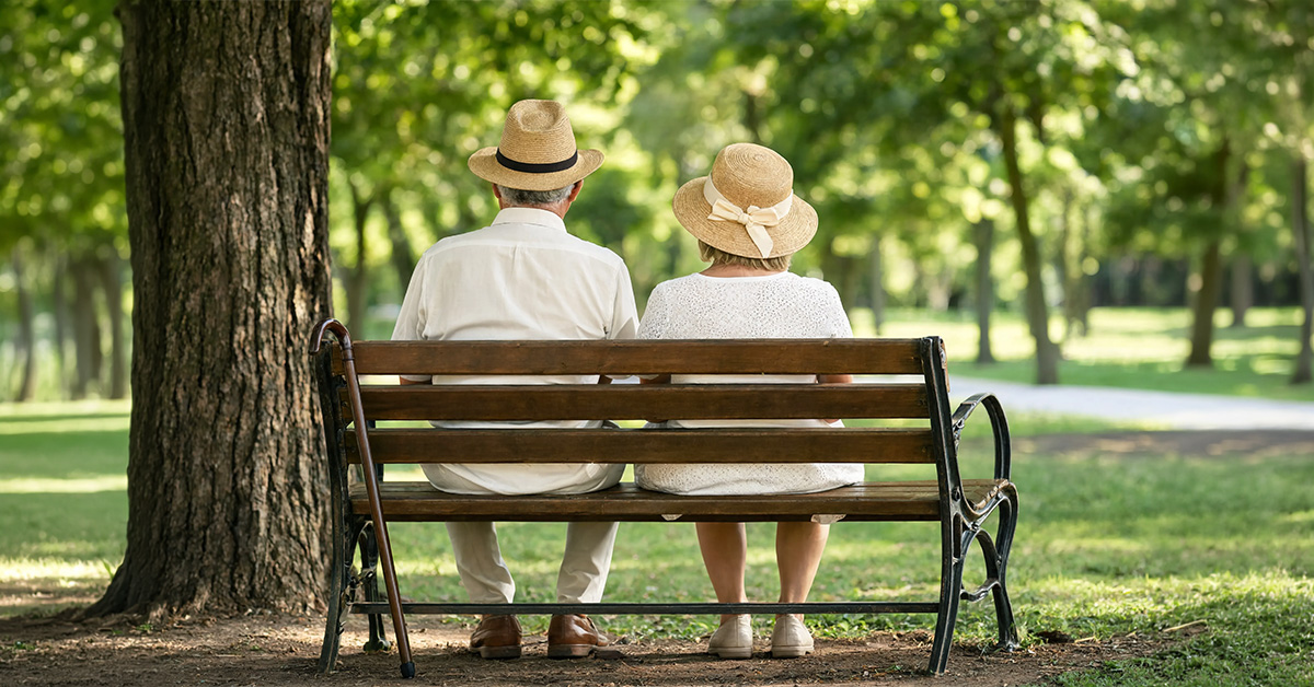Ein Seniorenpärchen sitzt gemeinsam auf einer Bank im Grünen, sie tragen zum Sonnenschutz helle Kleidung und Sonnenhüte