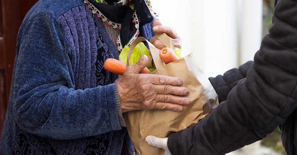 Eine ältere Person nimmt eine Einkaufstüte voller Lebensmittel von einer anderen Person entgegen.