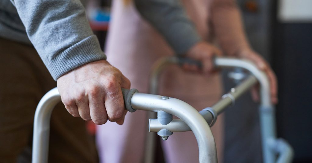 Eine ältere Person stützt sich an einem Rollator ab, im Hintergrund steht eine Pflegekraft zur Unterstützung bereit