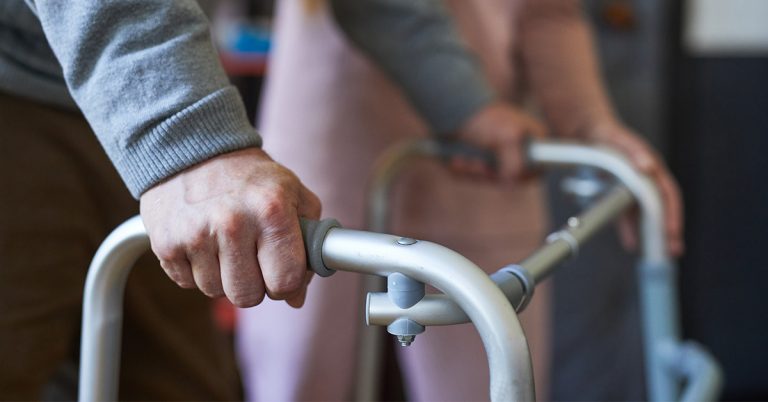 Eine ältere Person stützt sich an einem Rollator ab, im Hintergrund steht eine Pflegekraft zur Unterstützung bereit