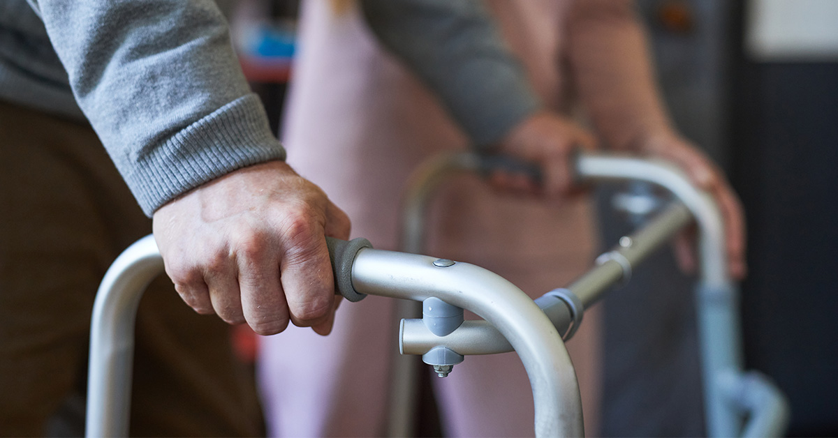 Eine ältere Person stützt sich an einem Rollator ab, im Hintergrund steht eine Pflegekraft zur Unterstützung bereit