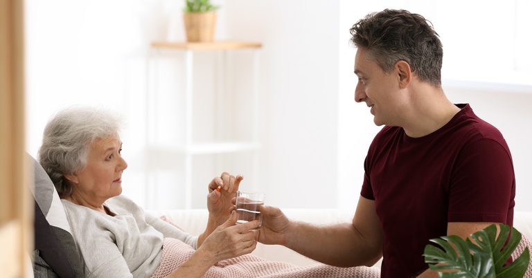 Eine ältere Dame liegt im Bett, ein Mann reicht ihr eine Tablette und ein Glas Wasser