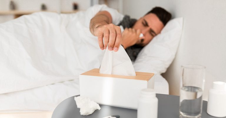 Ein junger Mann liegt krank im Bett und greift nach einem Taschentuch.